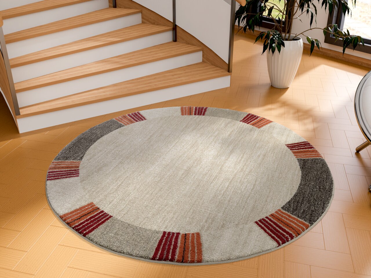 Runder Frisé-Teppich mit einem modernen Design in Beige, Grau und Rottönen, auf einem Holzfußboden vor einer Treppe platziert. Perspektive von oben.