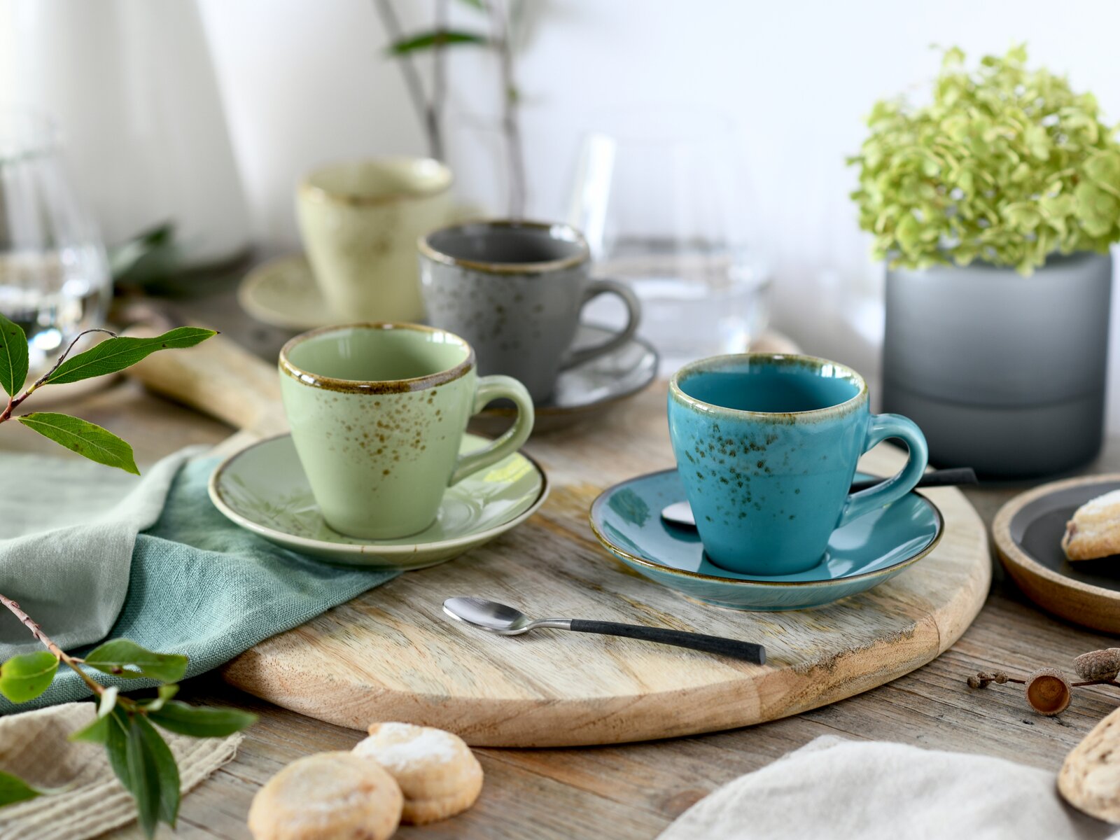 Rustikales Keramik-Kaffeetassen-Set mit Untertassen in Türkis und Hellgrün auf Holzbrett, perspektivisch in leichter Schrägansicht von vorn aufgenommen; im Hintergrund weitere Tassen und Deko.