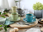 Rustikales Keramik-Kaffeetassen-Set mit Untertassen in Türkis und Hellgrün auf Holzbrett, perspektivisch in leichter Schrägansicht von vorn aufgenommen; im Hintergrund weitere Tassen und Deko.