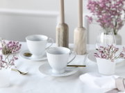 Zwei weiße geriffelte Porzellan-Teetassen mit Untertassen und goldenen Löffeln auf weiß gedecktem Tisch, mit zarten rosa Blumen und Kerzenständern im Hintergrund, Perspektive: leicht seitliche Ansicht auf Tischhöhe.