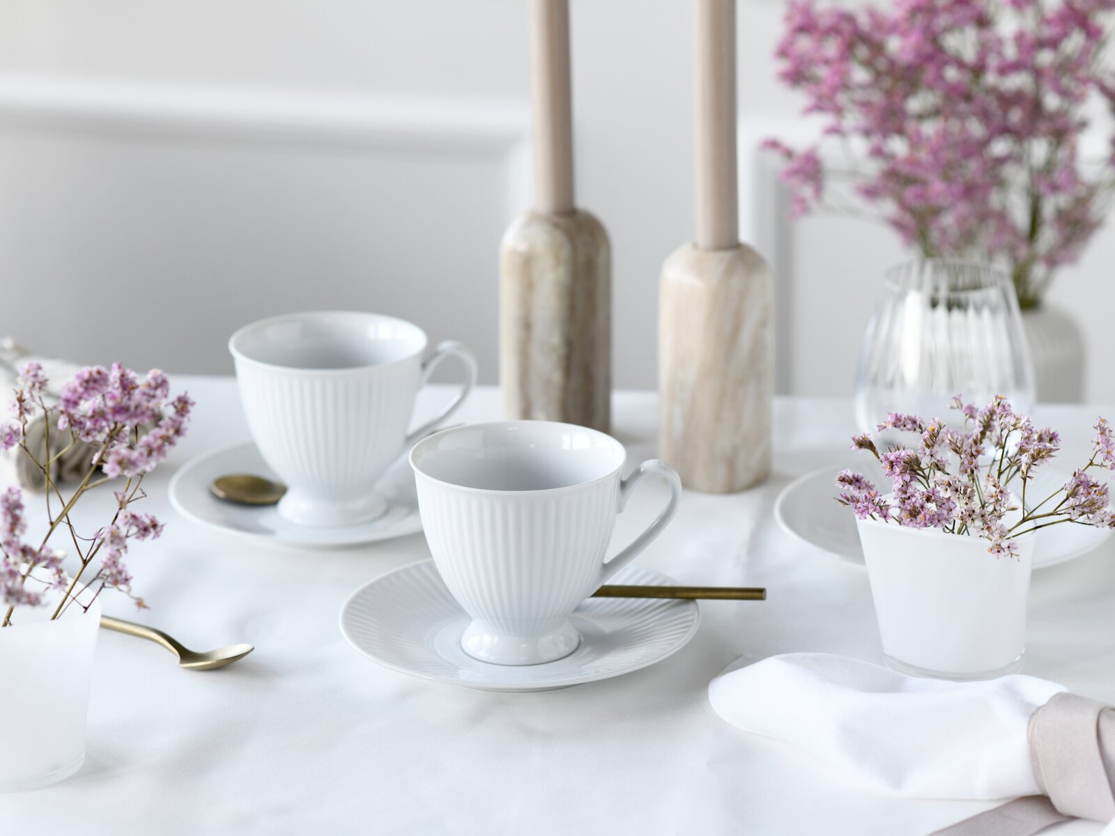Zwei weiße geriffelte Porzellan-Teetassen mit Untertassen und goldenen Löffeln auf weiß gedecktem Tisch, mit zarten rosa Blumen und Kerzenständern im Hintergrund, Perspektive: leicht seitliche Ansicht auf Tischhöhe.