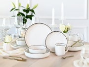 Elegantes weißes Geschirrset mit feinem dunklem Rand (Teller, Schalen und Becher) auf gedecktem Esstisch mit Gläsern und weißen Blumen, fotografiert in leicht erhöhter Frontperspektive.