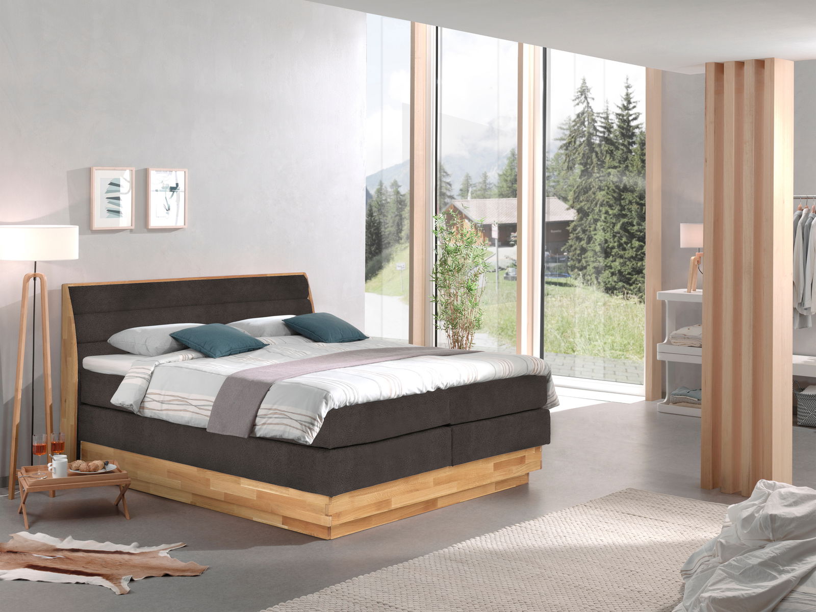Modernes Boxspringbett in einem hellen Schlafzimmer mit großen Fenstern und Blick auf die Natur, seitliche Perspektive.