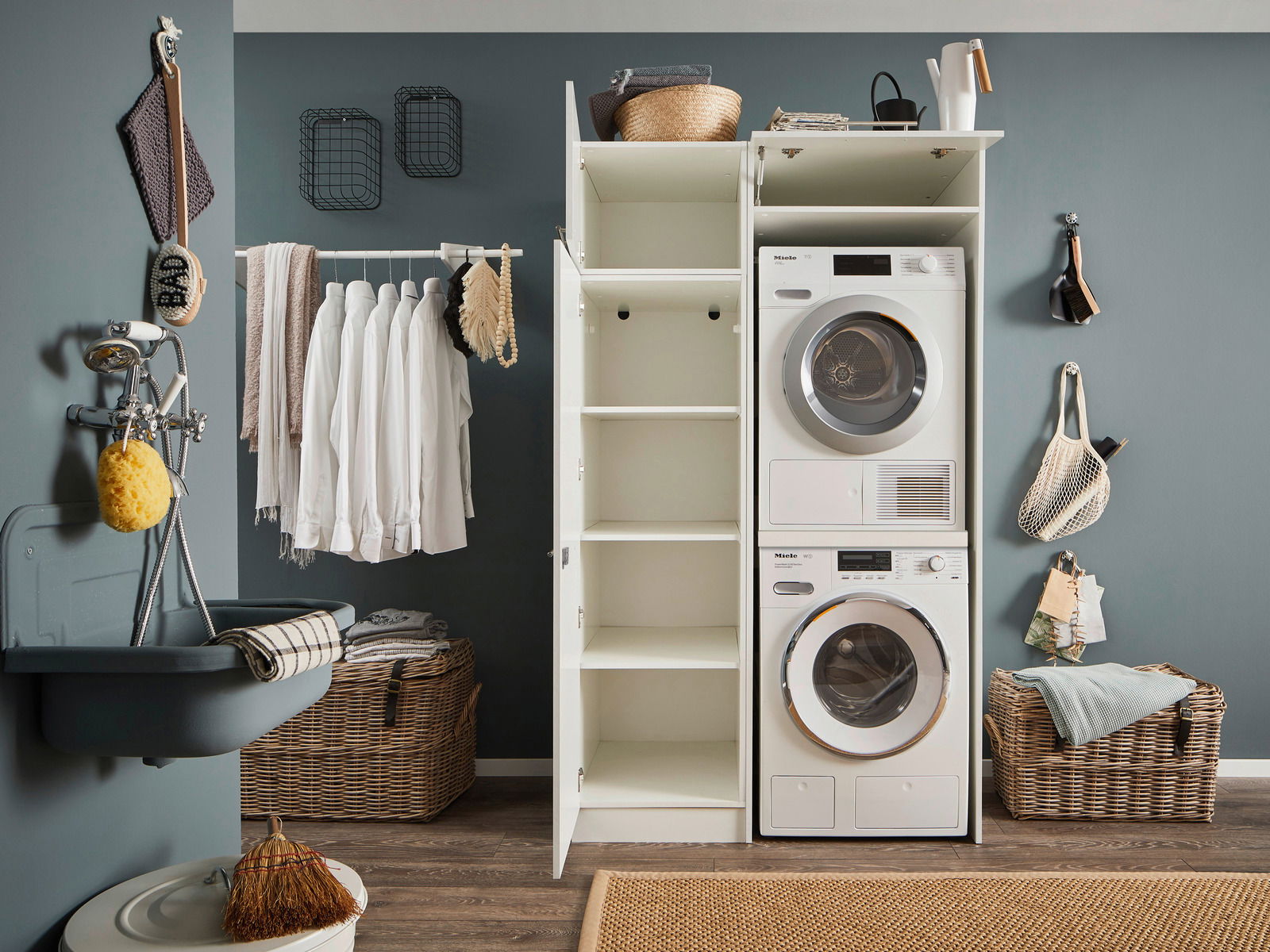 Hauswirtschaftsraum mit Waschmaschine und Trockner in einem Hochschrank, offene Regale, Wäschekorb und Waschbecken, seitliche Perspektive