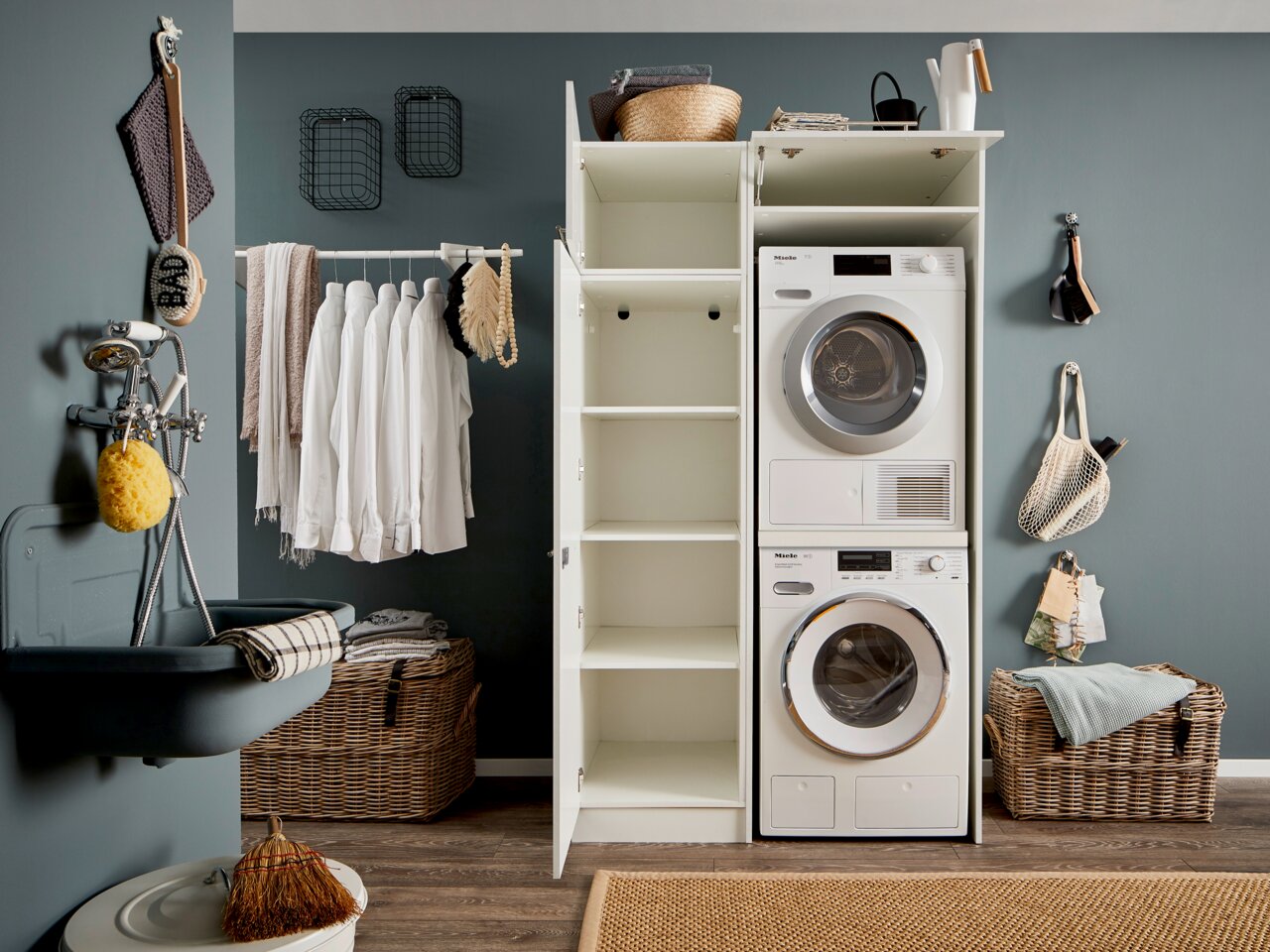 Hauswirtschaftsraum mit Waschmaschine und Trockner in einem Hochschrank, offene Regale, Wäschekorb und Waschbecken, seitliche Perspektive
