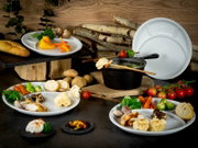Weißes, rundes Mehrkammer-Menüteller-Set in Food-Styling mit Gemüse und Brot sowie schwarzem Topf im Vordergrund; Perspektive: leicht von oben, frontal/3-4-Ansicht.