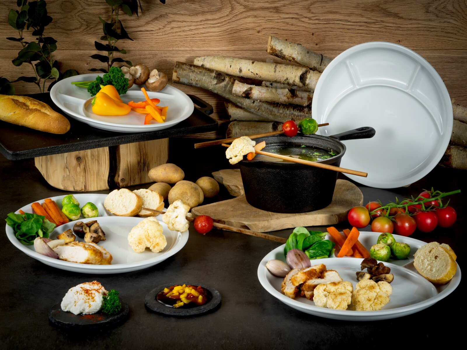 Weißes, rundes Mehrkammer-Menüteller-Set in Food-Styling mit Gemüse und Brot sowie schwarzem Topf im Vordergrund; Perspektive: leicht von oben, frontal/3-4-Ansicht.