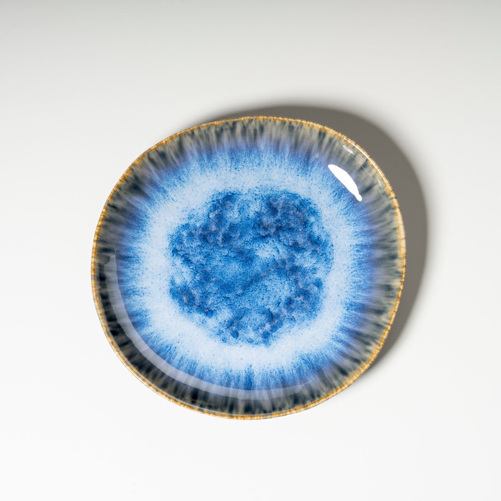 Runder, blau glasierter Keramik-Teller mit reaktivem Farbverlauf und braunem Rand, fotografiert in Draufsicht von oben auf weißem Hintergrund.