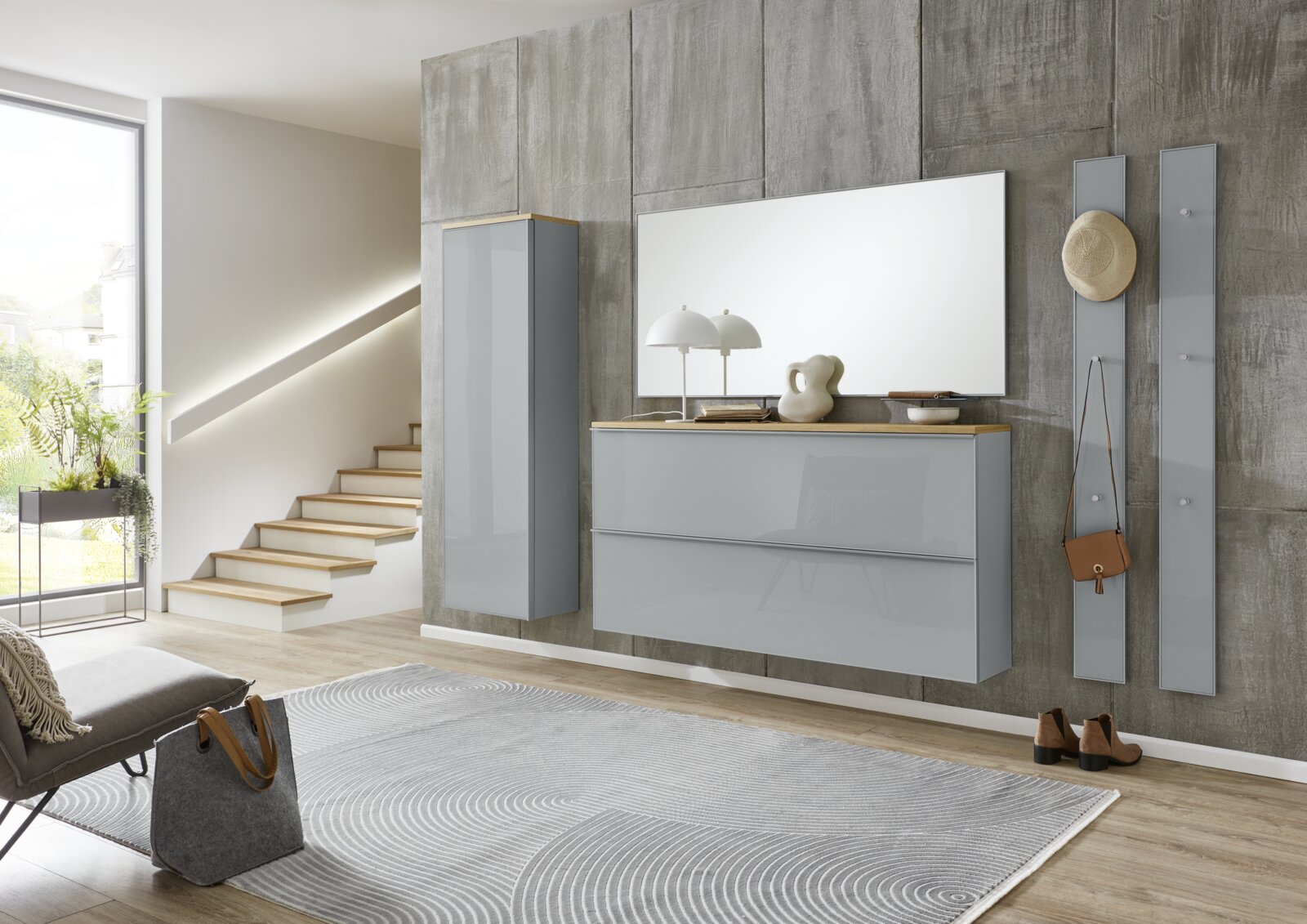 Modernes Flur-Set mit grauen Hochglanz-Wandmöbeln, großem Spiegel und Garderobenpaneelen an Betonoptik-Wand, frontal aus leicht seitlicher Perspektive aufgenommen.