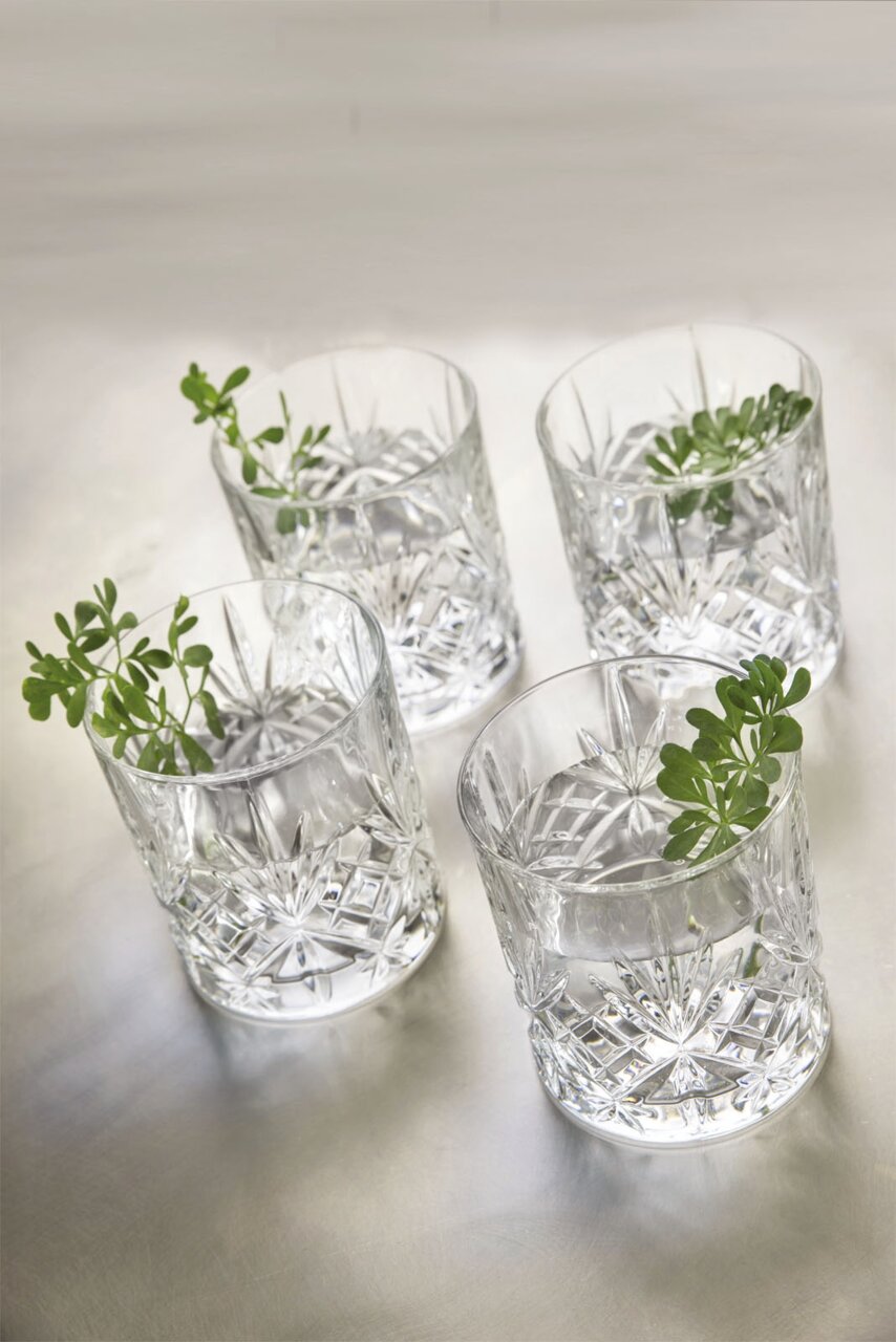 Vier kristalheldere whiskyglazen met decoratieve slijpvorm, gefotografeerd van bovenaf, met kleine groene plantjes als versiering.