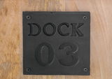 Nahaufnahme einer schwarzen Metallplatte mit der Aufschrift 'DOCK 03' auf einer Holzoberfläche.
