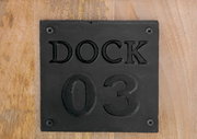 Nahaufnahme einer schwarzen Metallplatte mit der Aufschrift 'DOCK 03' auf einer Holzoberfläche.