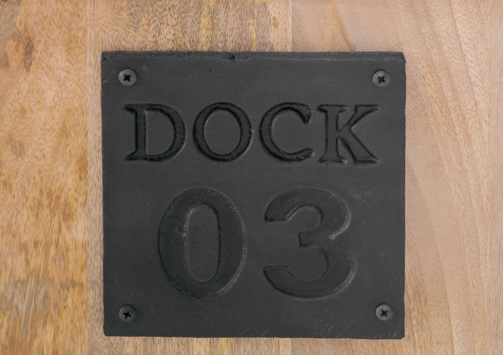 Nahaufnahme einer schwarzen Metallplatte mit der Aufschrift 'DOCK 03' auf einer Holzoberfläche.