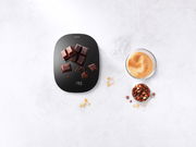 Digitale Küchenwaage in Schwarz mit Schokoladenstücken darauf, aus der Vogelperspektive fotografiert.