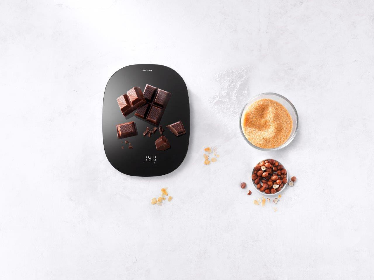 Digitale Küchenwaage in Schwarz mit Schokoladenstücken darauf, aus der Vogelperspektive fotografiert.