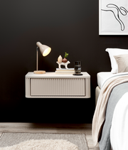 Schwebender, cremefarbener Nachttisch mit geriffelter Front und abgerundeten Kanten neben einem Bett, darauf Tischlampe, Bücher und Deko – Frontansicht in leichter Seitenperspektive.