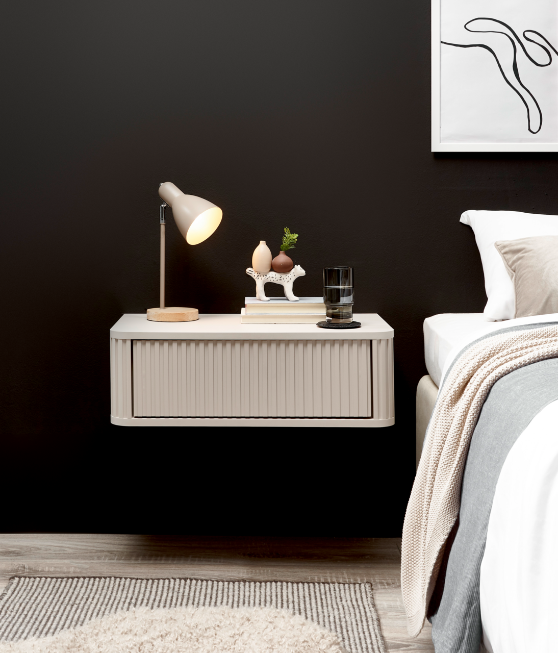 Schwebender, cremefarbener Nachttisch mit geriffelter Front und abgerundeten Kanten neben einem Bett, darauf Tischlampe, Bücher und Deko – Frontansicht in leichter Seitenperspektive.