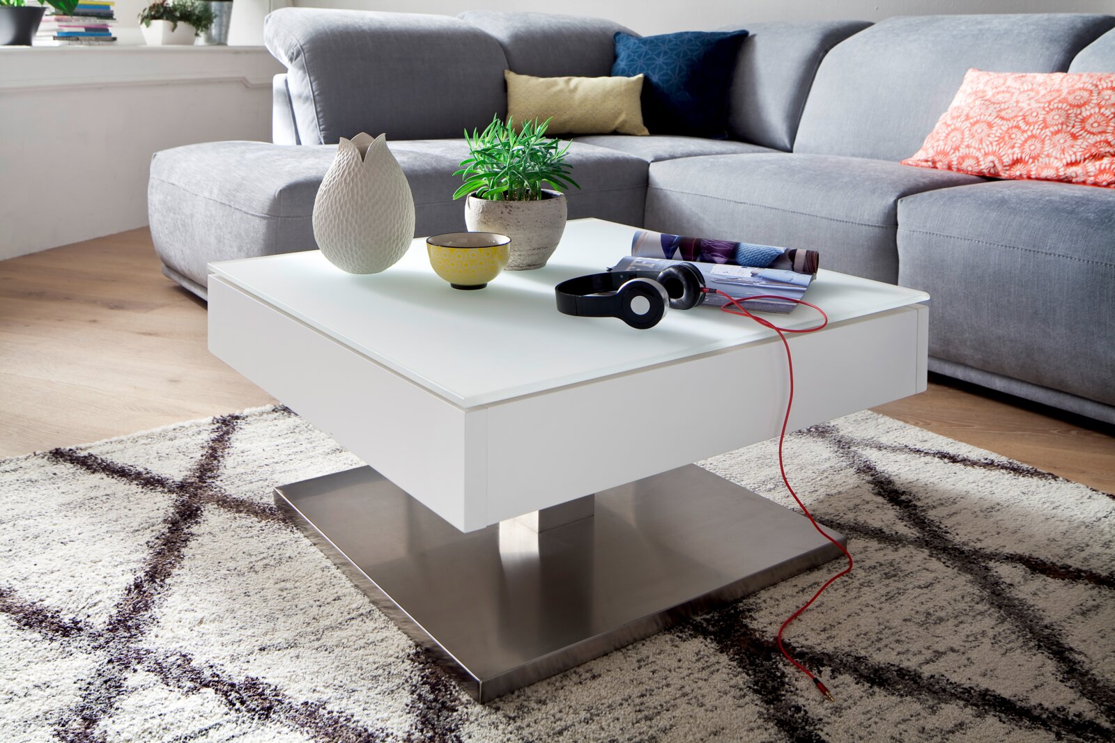 Zurbrüggen Couchtisch MARIKO Moderner weißer Couchtisch mit quadratischer Glasplatte und Metallfuß, dekoriert mit einer Vase, einer Pflanze, einer Schale und Kopfhörern, in einem gemütlichen Wohnzimmer mit grauem Sofa, aus seitlicher Perspektive.