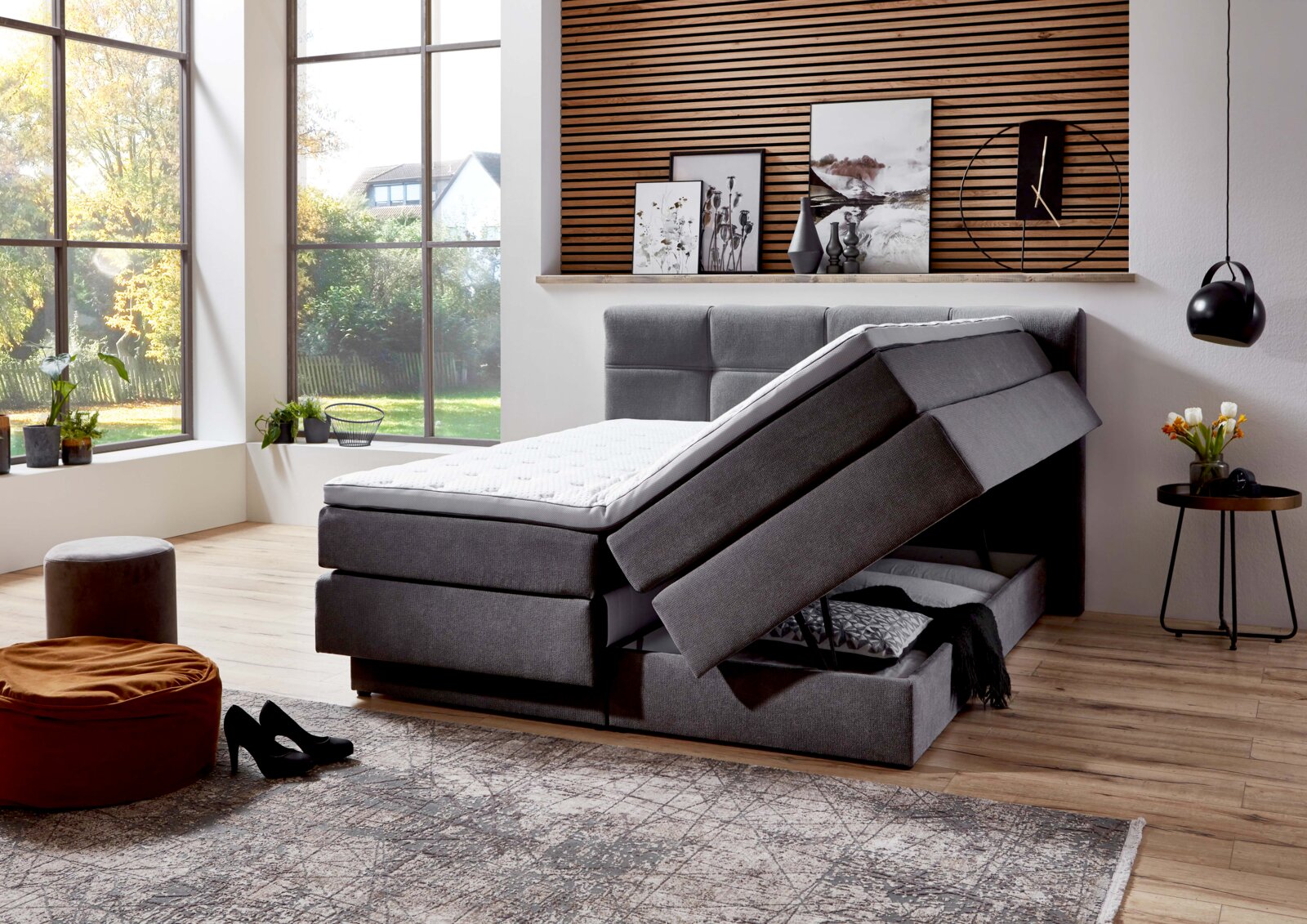 Graues Boxspringbett mit Stauraumfunktion, seitliche Perspektive, in modernem Schlafzimmer mit großen Fenstern und stilvoller Dekoration.