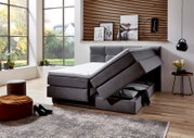 Graues Boxspringbett mit Stauraumfunktion, seitliche Perspektive, in modernem Schlafzimmer mit großen Fenstern und stilvoller Dekoration.
