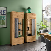 Zwei moderne Highboards aus Holz mit Glaseinsätzen, dekoriert mit Vasen und Geschirr, in einem stilvoll eingerichteten Raum mit grüner Wand und großem Fenster. Perspektive von vorne.