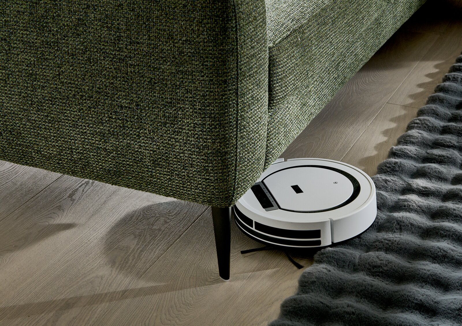 Ein grünes Longchair-Sofa mit einem Saugroboter, der darunter reinigt. Perspektive von der Seite.