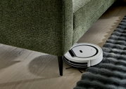Ein grünes Longchair-Sofa mit einem Saugroboter, der darunter reinigt. Perspektive von der Seite.