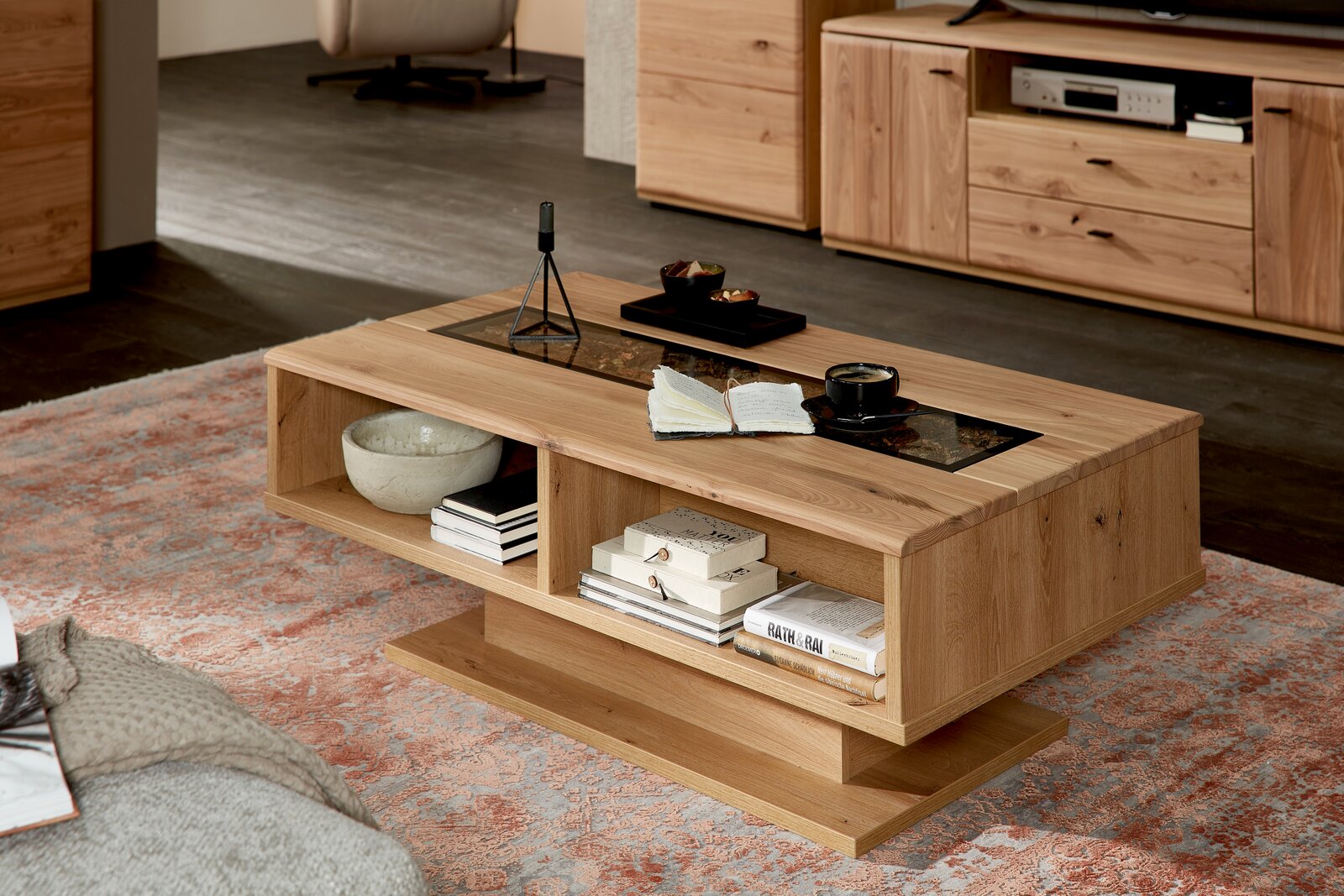 Moderner Couchtisch aus Holz mit offenen Fächern, dekoriert mit Büchern und einer Tasse Kaffee, in einem gemütlichen Wohnzimmer. Perspektive von schräg vorne.