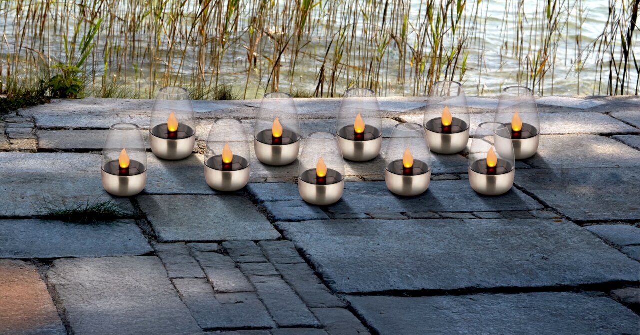 Ambia Garden Solarleuchten 10er-Set Mehrere LED-Teelichter in Glas-Windlichtern mit Edelstahlfuß auf Steinplatten am Seeufer, seitliche Perspektive auf die Gruppe