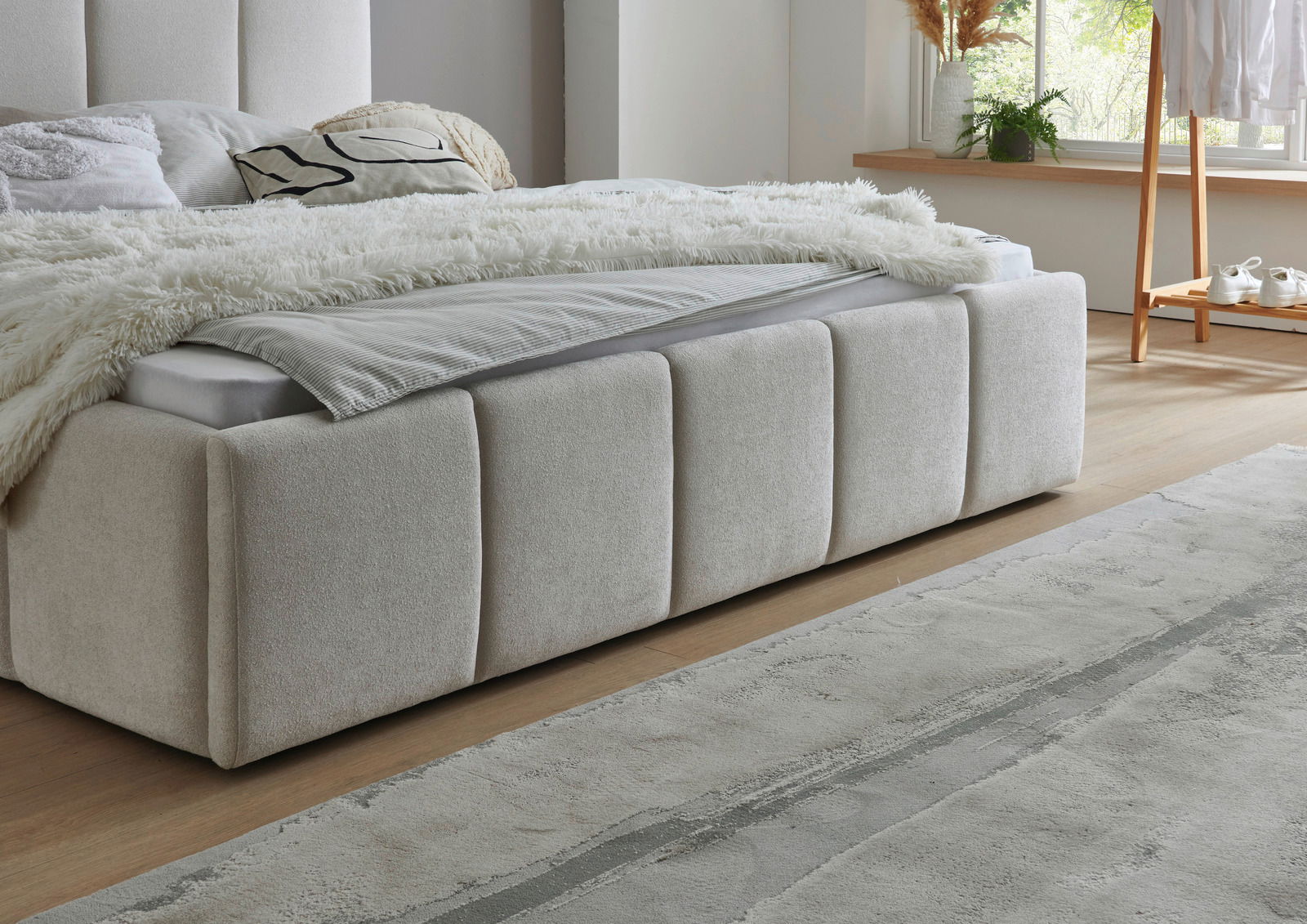 Seitliche Perspektive auf ein elegantes Polsterbett mit beigem Stoffbezug, dekoriert mit Kissen und einer flauschigen Decke, in einem hellen, modernen Schlafzimmer.