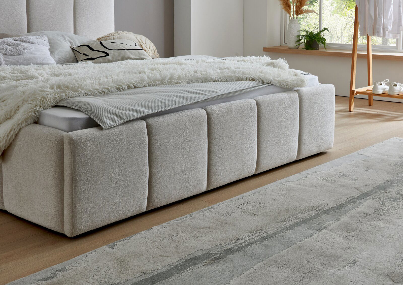 Seitliche Perspektive auf ein elegantes Polsterbett mit beigem Stoffbezug, dekoriert mit Kissen und einer flauschigen Decke, in einem hellen, modernen Schlafzimmer.