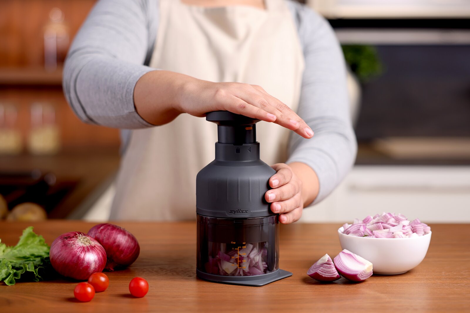 Vooraanzicht op een handmatige uienhakker op het aanrecht, bediend met een drukmechanisme, met rode uien en cherrytomaten ernaast en een kom met gesnipperde ui.