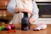 Vooraanzicht op een handmatige uienhakker op het aanrecht, bediend met een drukmechanisme, met rode uien en cherrytomaten ernaast en een kom met gesnipperde ui.