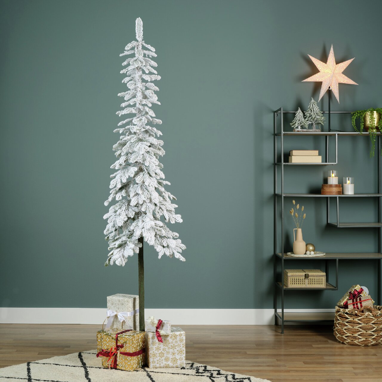Schneebedeckte künstliche Tanne in einem modernen Wohnzimmer, frontal fotografiert, mit Geschenken am Boden und einem Regal im Hintergrund.