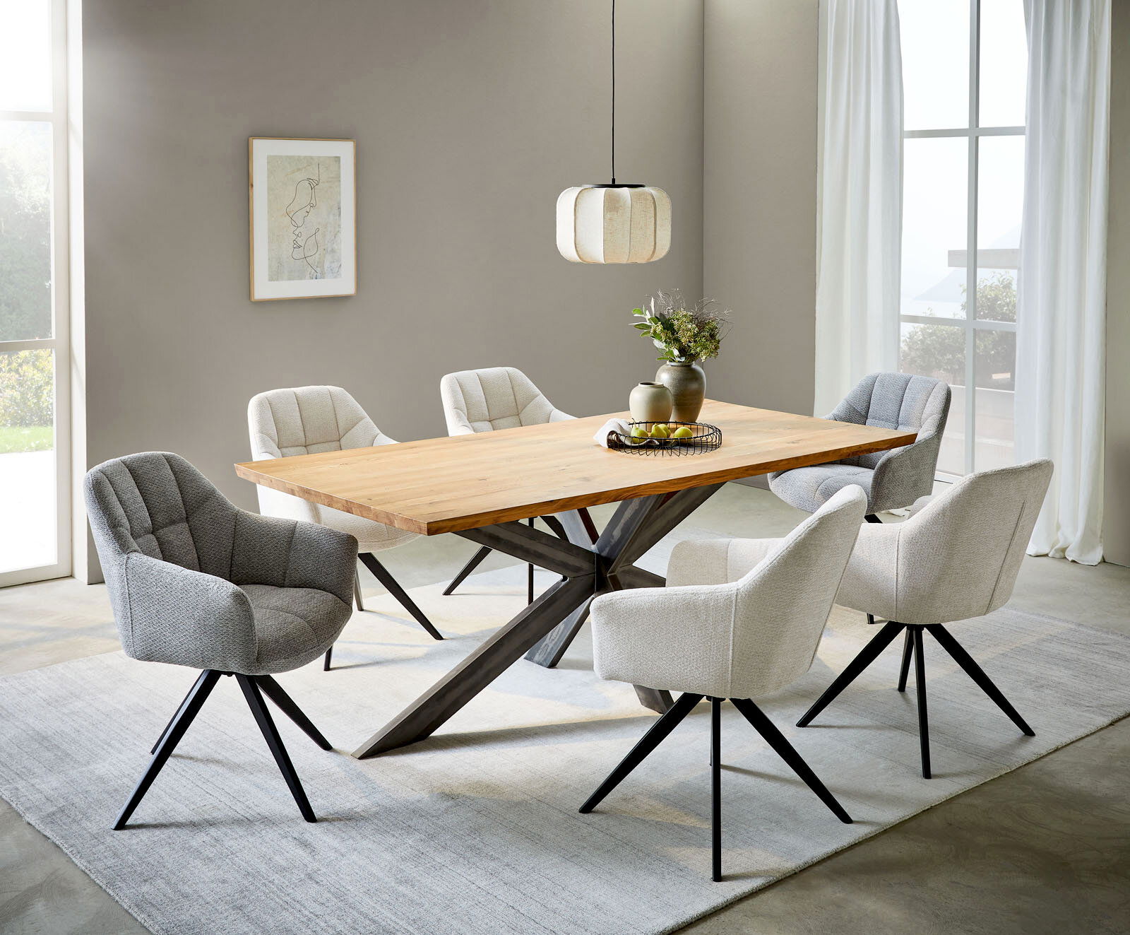 Moderner Esstisch mit heller Holzplatte und schwarzem X-Gestell, umgeben von gepolsterten Stühlen in Grau und Beige in einem hellen Esszimmer; Perspektive: leicht erhöhte Frontansicht schräg von links.