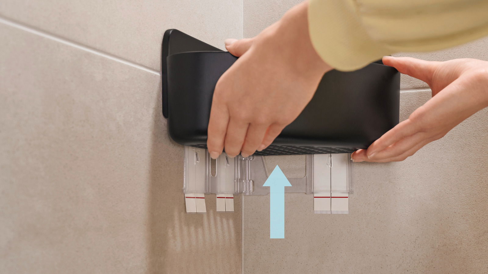 Nahaufnahme eines schwarzen Eckduschkorbs, der an einer Fliesenwand montiert wird. Hände justieren den Korb, während ein Pfeil die Richtung des Anbringens anzeigt.