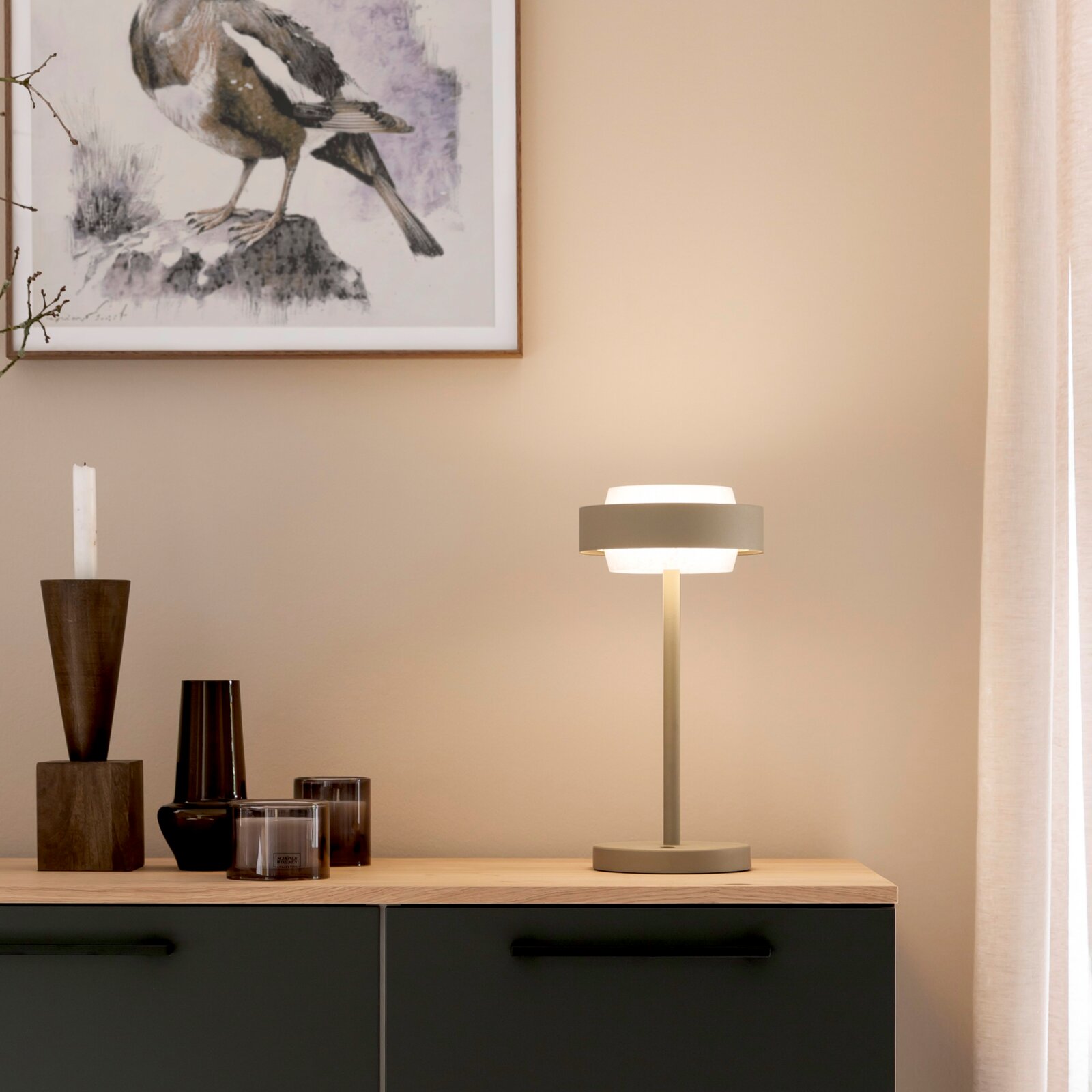 Minimalistische Tischlampe in Beige mit rundem Standfuß und doppellagigem Schirm, eingeschaltet auf einem Sideboard im Wohnzimmer, frontal aus leicht seitlicher Perspektive fotografiert.