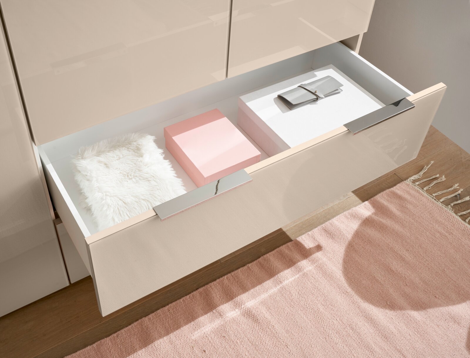 Geöffnete Schublade eines beigen Schranks mit Chromgriffen, darin rosa und weiße Boxen, ein weißes Fellkissen und ein graues Etui; Perspektive: schräg von oben