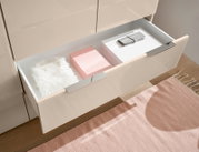 Geöffnete Schublade eines beigen Schranks mit Chromgriffen, darin rosa und weiße Boxen, ein weißes Fellkissen und ein graues Etui; Perspektive: schräg von oben
