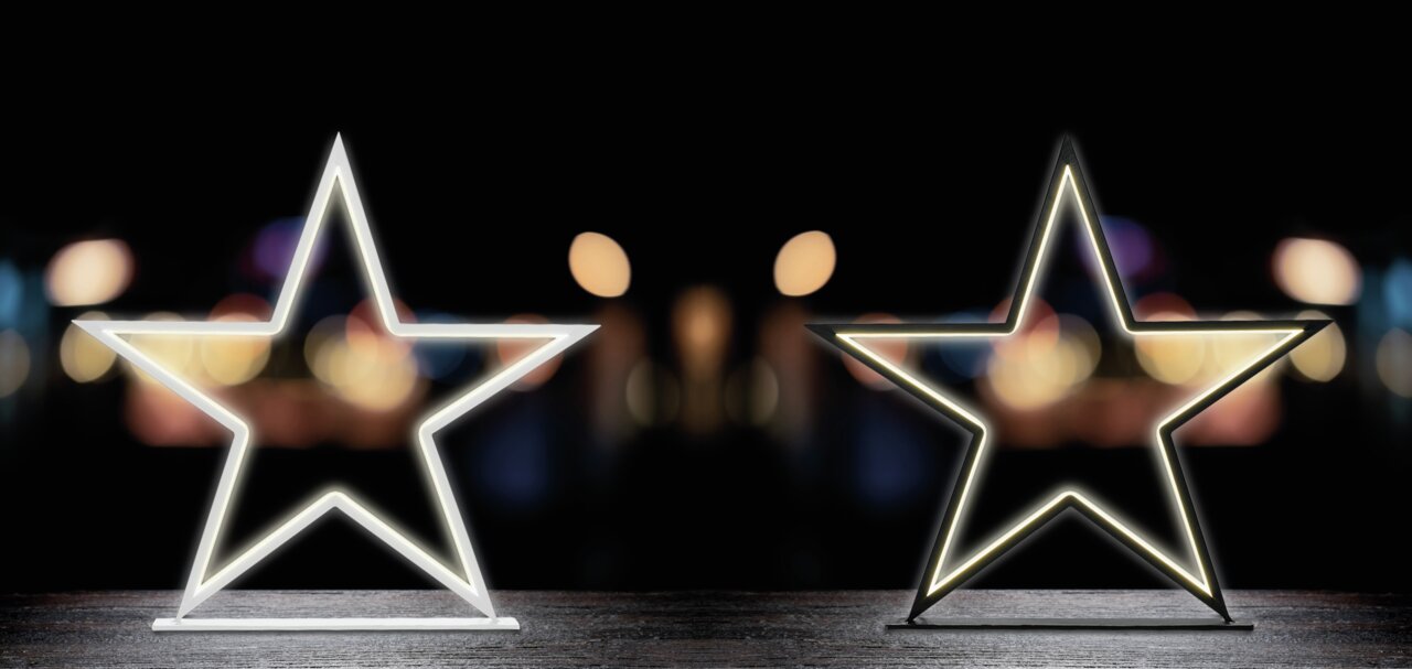 Zwei leuchtende LED-Sterne auf einem Tisch vor einem unscharfen, dunklen Hintergrund. Frontalansicht.