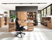 Moderne Büromöbel in einem hellen Raum mit großen Fenstern, bestehend aus einem Schreibtisch, Rollcontainer, Aktenschränken und Regalen aus Holz. Perspektive von vorne.