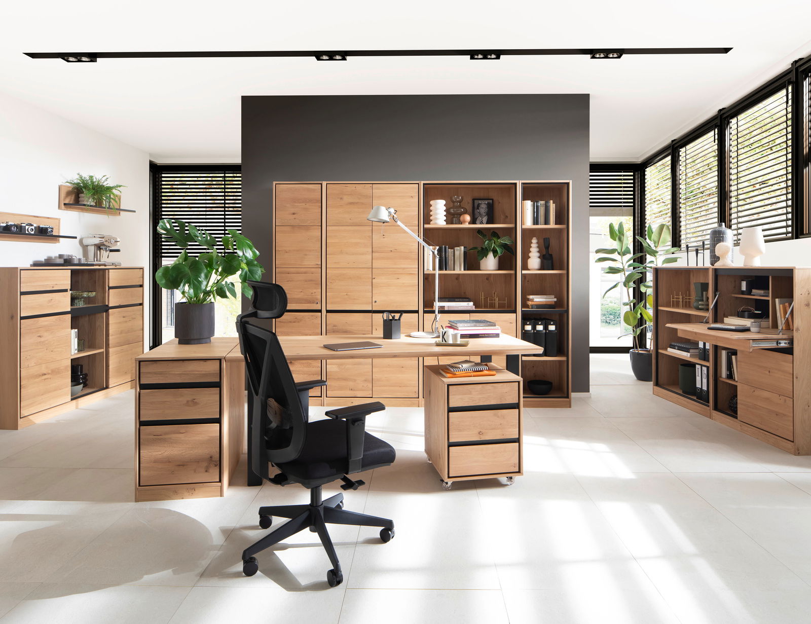 Moderne Büromöbel in einem hellen Raum mit großen Fenstern, bestehend aus einem Schreibtisch, Rollcontainer, Aktenschränken und Regalen aus Holz. Perspektive von vorne.