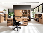 Moderne Büromöbel in einem hellen Raum mit großen Fenstern, bestehend aus einem Schreibtisch, Rollcontainer, Aktenschränken und Regalen aus Holz. Perspektive von vorne.