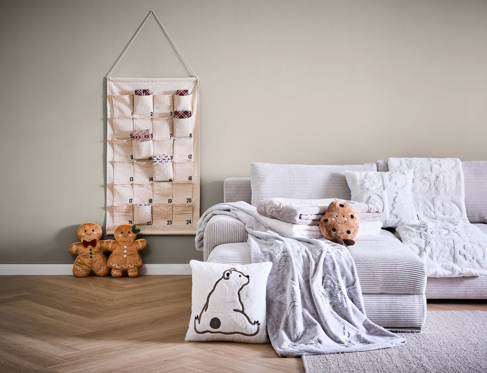 Gemütliche Wohnzimmer-Szene mit einer weichen, hellen Wohndecke auf einem Sofa, dekoriert mit einem Kissen und einem Plüschkeks. Im Hintergrund hängt ein Adventskalender an der Wand. Perspektive: Frontalansicht.
