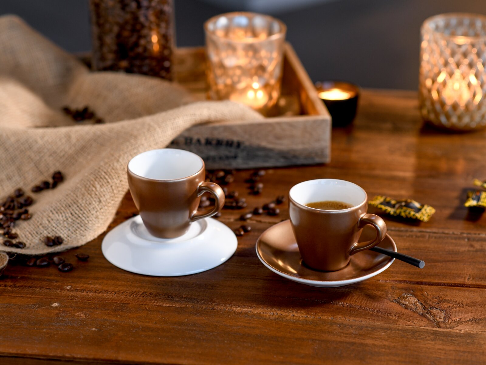 Creatable Espressoset 4-tlg GOLDEN DAYS Zwei bronzefarbene Espressotassen mit Untertassen auf rustikalem Holztisch, umgeben von Kaffeebohnen und Kerzenlicht, aufgenommen aus leicht erhöhter Perspektive schräg von vorn