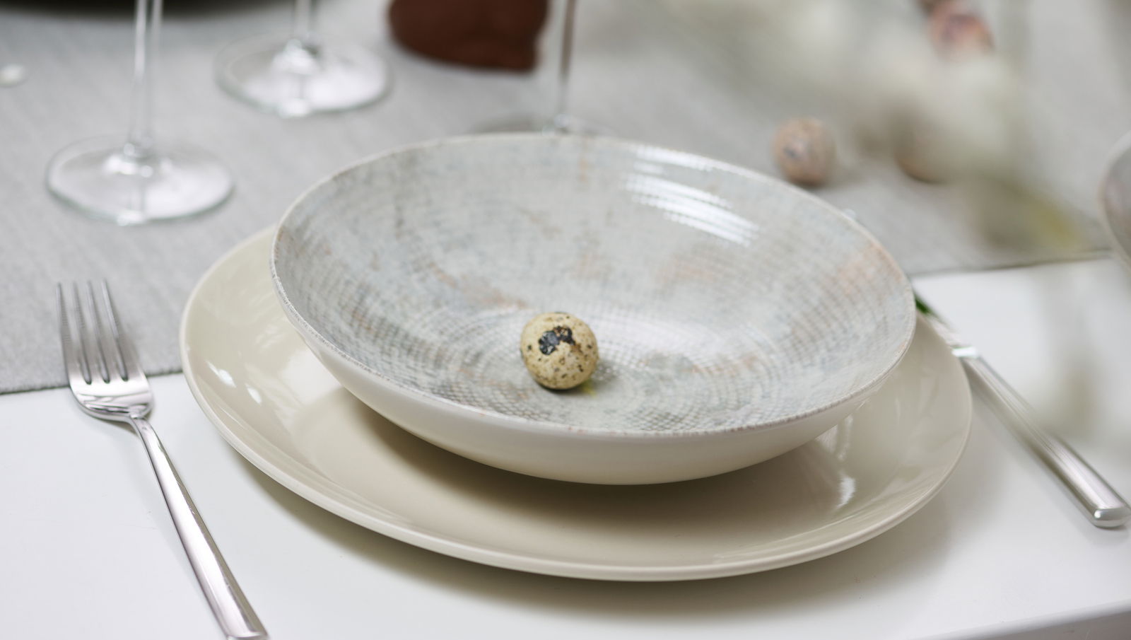Ritzenhoff & Breker Dessertteller CECINA beige Ein gedeckter Tisch mit einem flachen Teller und einer Schale darauf, aus der Vogelperspektive fotografiert. In der Schale liegt ein Wachtelei. Besteck und Weingläser sind ebenfalls zu sehen.