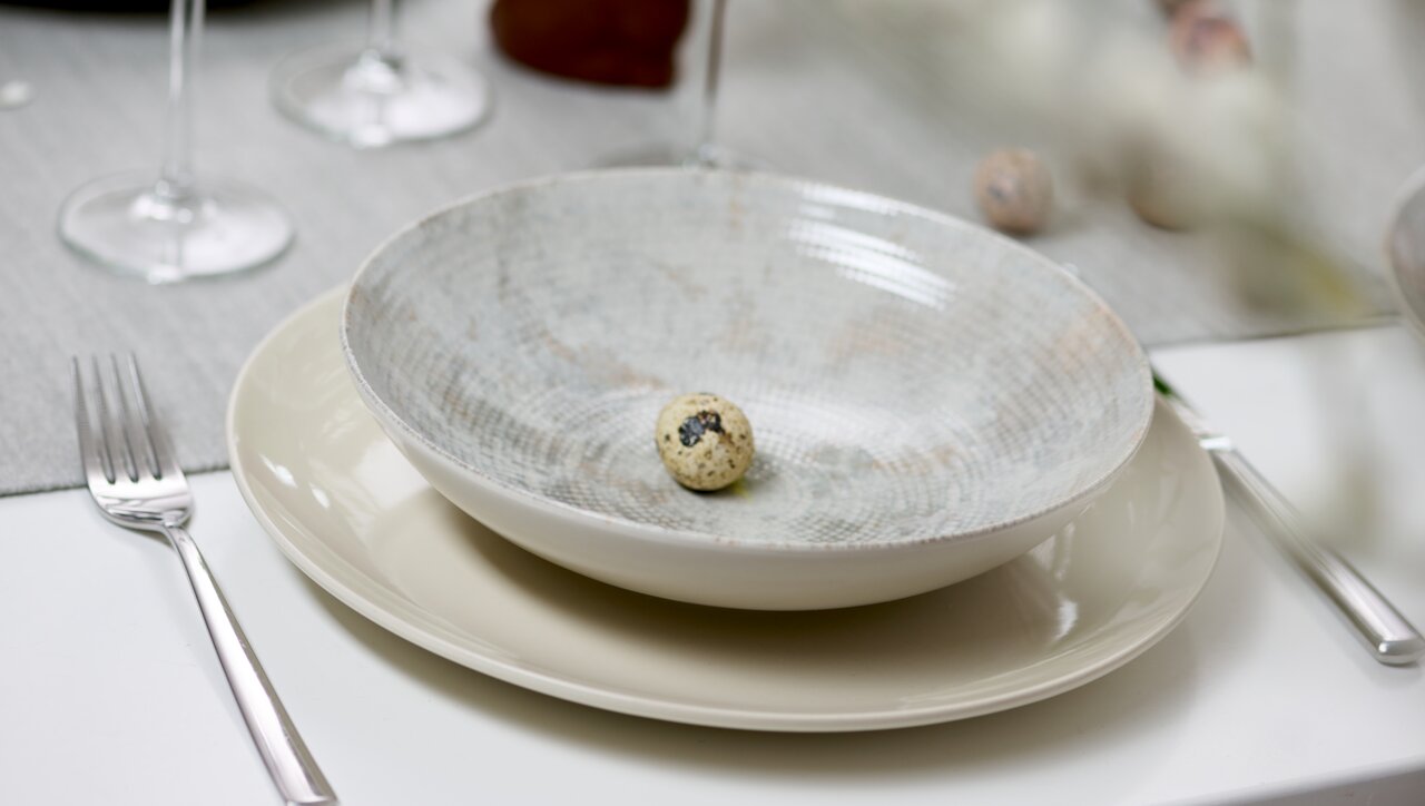 Ritzenhoff & Breker Dessertteller CECINA beige Ein gedeckter Tisch mit einem flachen Teller und einer Schale darauf, aus der Vogelperspektive fotografiert. In der Schale liegt ein Wachtelei. Besteck und Weingläser sind ebenfalls zu sehen.