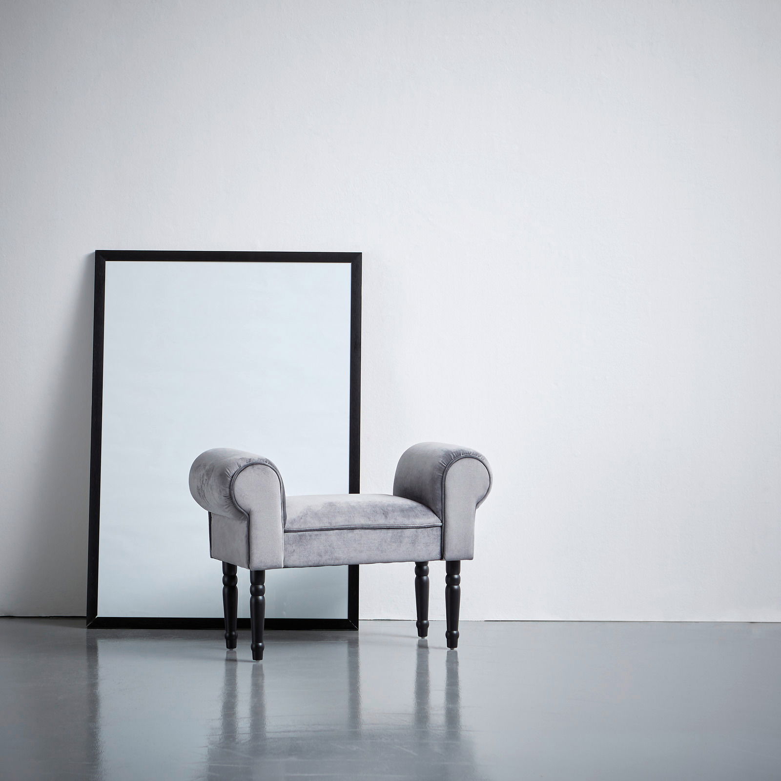 XORA Hocker LORE Grauer Hocker mit gepolsterten Armlehnen und schwarzen Beinen, seitliche Perspektive, vor einem großen Spiegel aufgestellt.