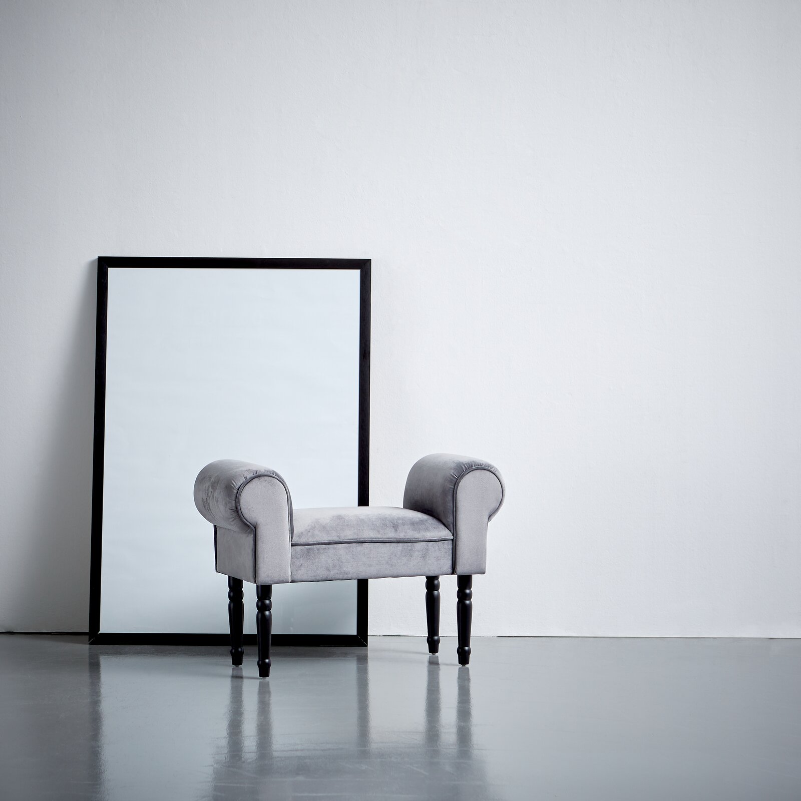 Grauer Hocker mit gepolsterten Armlehnen und schwarzen Beinen, seitliche Perspektive, vor einem großen Spiegel aufgestellt.