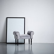 Grauer Hocker mit gepolsterten Armlehnen und schwarzen Beinen, seitliche Perspektive, vor einem großen Spiegel aufgestellt.